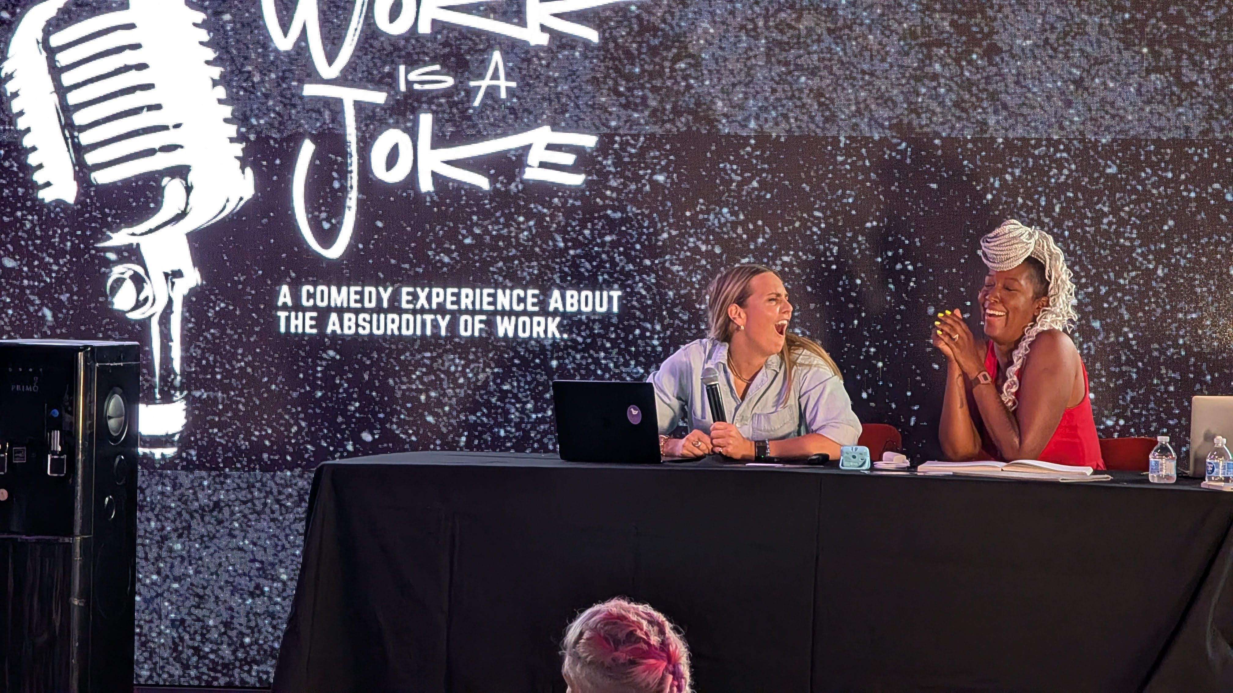 Casey Boyles co-hosting the Work is a Joke comedy show onstage with a fellow host, both laughing behind a table, with the show's title projected on the screen behind them